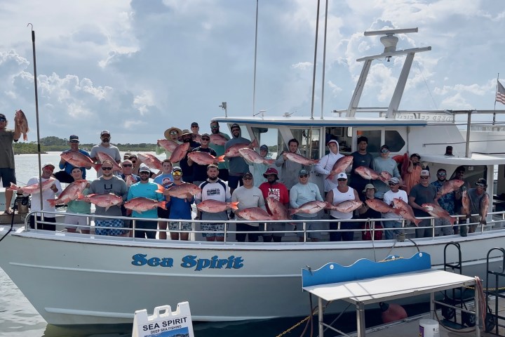 Group of people on a boat holding large fish, under cloudy sky.